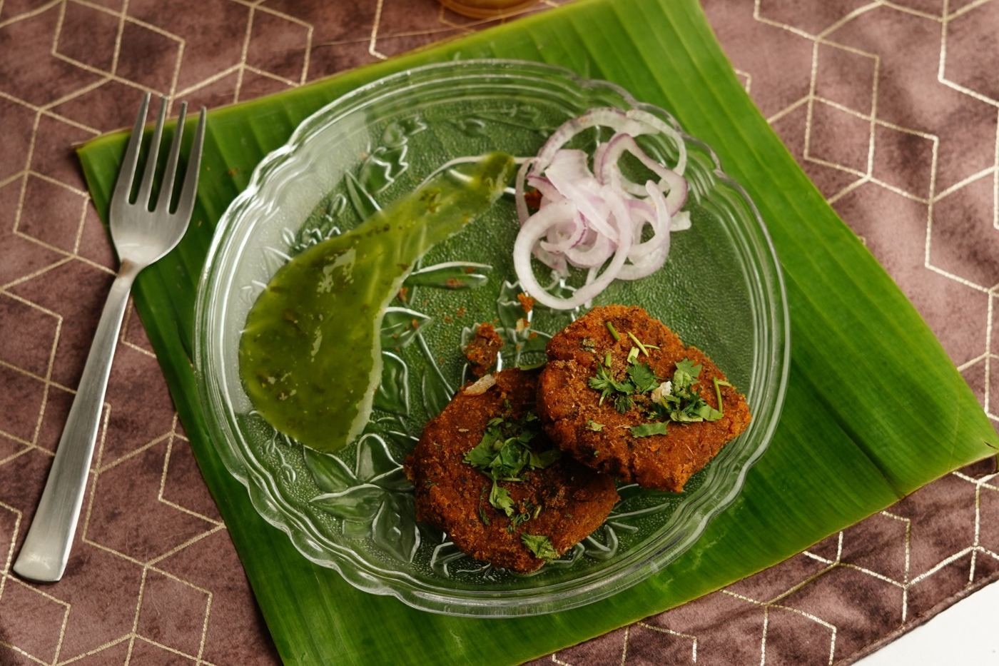 Two veg shami kababs with green chutney and onion rings on a banana leaf